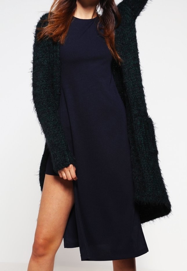 Femme portant une robe noire sans manches avec une fente haute et un long cardigan texturé vert foncé, posant avec un bras levé.