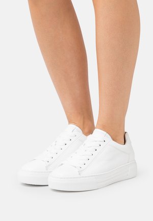 Jambes nues portant des baskets blanches basses à lacets avec des semelles épaisses en caoutchouc, présentées sur un fond blanc uni.