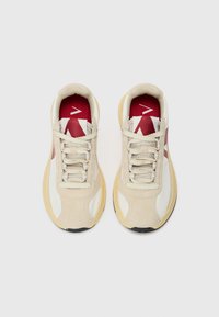 Beige athletic shoes with red accents, featuring a mix of suede and textile material, round toe, and textured sole. Laces are light gray.