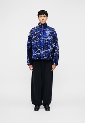 Vīrietis stāv ar seju pret priekšu, valkājot zilu rakstainu polarvates jaku ar rāvējslēdzēju, platās melnās bikses un melnus apavus uz vienkārša gaiša fona.