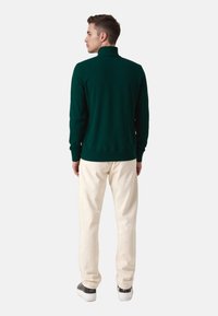 Grüner Sweatshirt mit hohem Kragen und langen Ärmeln, kombiniert mit cremefarbener Hose und schwarzen Sneakers, die von hinten ein sauberes, minimalistisches Design zeigen.