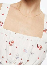 Robe florale blanche avec des motifs rouges et violets, présentant une encolure carrée et une texture froncée. Un collier en chaîne dorée accentue l'encolure.