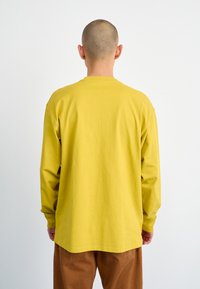 Long-sleeve t-shirt in mustard yellow cotton, features a round neckline, and relaxed fit, paired with brown trousers, viewed from the back.