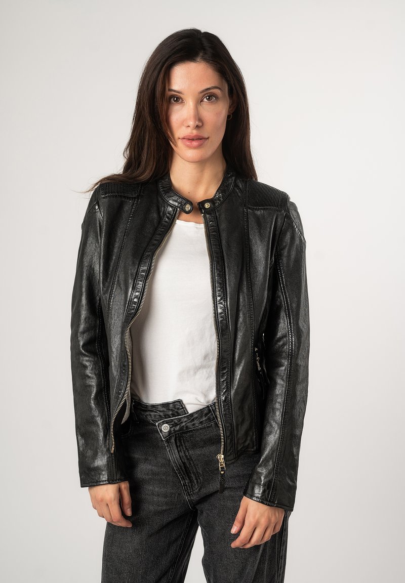 Schwarze Lederjacke mit einem Reißverschluss vorne, rundem Kragen und strukturierten Schultern, kombiniert mit dunkelgrauen Jeans und einem weißen Hemd darunter.