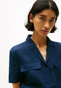 Camicia blu navy a maniche corte con patta con bottoni, due tasche sul petto e colletto strutturato. Tessuto liscio con una leggera lucentezza.