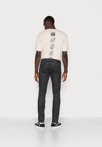 T-shirt gráfica bege com ilustrações nas costas, combinada com jeans escuros de corte justo e ténis pretos com detalhes brancos.
