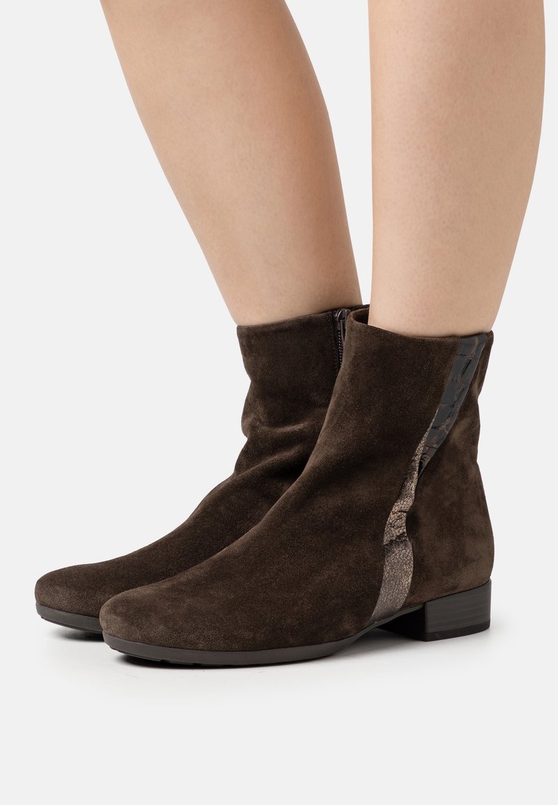 Bottines en daim marron foncé avec un petit talon et une fermeture éclair latérale portées sur des jambes nues, sur un fond uni.