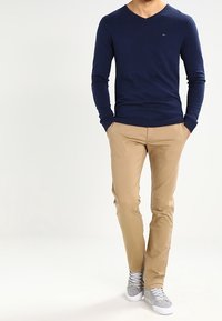 Tmavě modrý svetr do V, khaki úzké chino kalhoty a šedé tenisky. Svetr má malé logo na hrudi; chino kalhoty mají hladkou texturu.