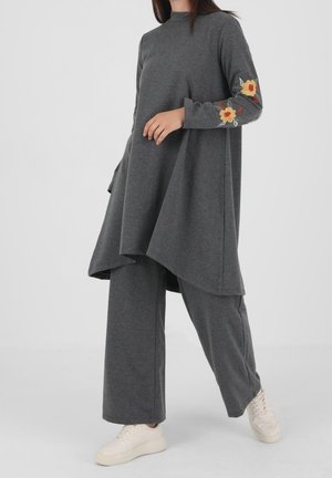Personne portant une tenue ample gris foncé avec un long haut présentant une broderie de fleurs jaunes et orange sur les manches et un pantalon large assorti.