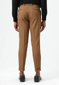 Bruine op maat gemaakte broek, lichtgewicht stof, slanke pasvorm, omgeslagen boorden, twee achterzakken en fijn gestikte details. Draagt bijpassende zwarte schoenen.