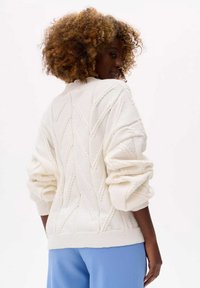 White knitted sweater with a diamond pattern, puffy sleeves, and a ribbed hem. Worn with light blue trousers.