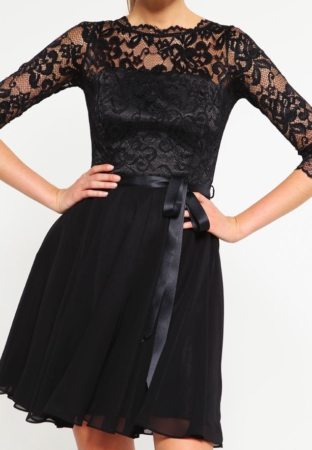 Robe noire avec manches longues en dentelle, corsage à motifs floraux, taille ajustée avec ruban en satin et jupe fluide. Mains sur les hanches.