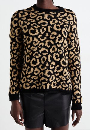 Person iført en langærmet, sort og tan leopardmønstret sweater kombineret med sorte lædershorts, stående med armene afslappet ned langs siden.