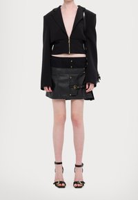 Svart kort jacka med dragkedja, tillsammans med en kjol i konstläder som har guldfärgad hardware, fickor och spänndetaljer. Svarta sandaletter med remmar.