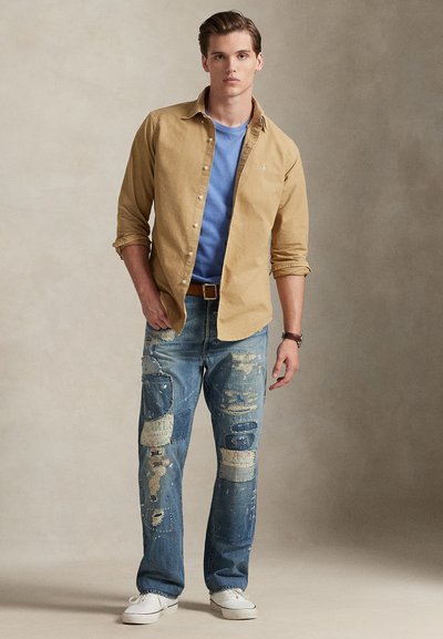 Μπεζ πουκάμισο, μπλε T-shirt από κάτω, φθαρμένα τζιν με ξεθωριασμένες περιοχές και λευκά αθλητικά παπούτσια. Απλό ρολόι στο χέρι.