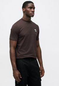 Άντρας που στέκεται φορώντας ένα σκούρο καφέ κοντομάνικο πουκάμισο με ένα μικρό λευκό και μαύρο λογότυπο στο στήθος και μαύρο παντελόνι μπροστά σε απλό λευκό φόντο.