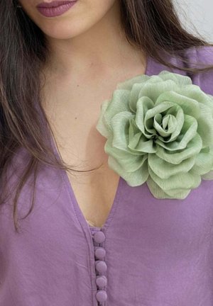 Vrouw draagt een paarse top met knopen en een grote lichtgroene stofbloem gespeld bij de halslijn, lang donker haar zichtbaar.