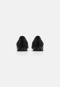 Zapatos negros tipo slip-on con parte superior texturizada y acentos lisos de charol negro en el talón. Presentan una forma redondeada y un perfil bajo.