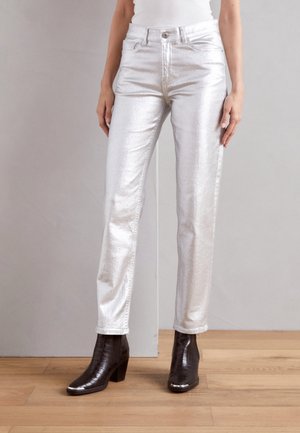 Personne portant un pantalon argenté brillant et des bottines noires à talons, debout sur un sol en bois clair contre un mur gris.