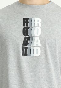 Camiseta gris de manga corta con un diseño de texto apilado en estilo 3D en negro, gris y blanco, con una cita impresa en la parte inferior derecha. Textura suave.