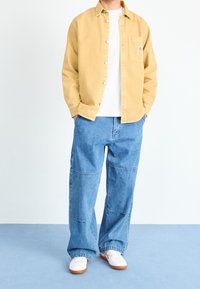 Ljusa gul skjorta med knappar över en vit t-shirt, kombinerad med löst sittande blå denimbyxor. Vita sneakers med bruna sulor fullbordar outfiten.