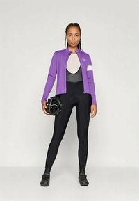 Paarse fietsjack met lange mouwen over een zwart-grijze aansluitende body, gecombineerd met zwarte leggings en een fietshelm in de hand.