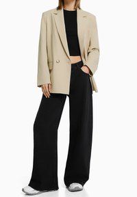 Beige oversized blazer met een enkele knoop, gecombineerd met een zwart cropped topje en wijde zwarte jeans. Witte sportschoenen maken de look compleet.