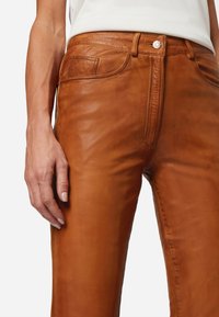 Braune Lederhosen mit einem taillierten Design, sichtbaren Nähten, Fronttaschen und einem Metallknopfverschluss. Glatte Textur mit einem leichten Glanz.