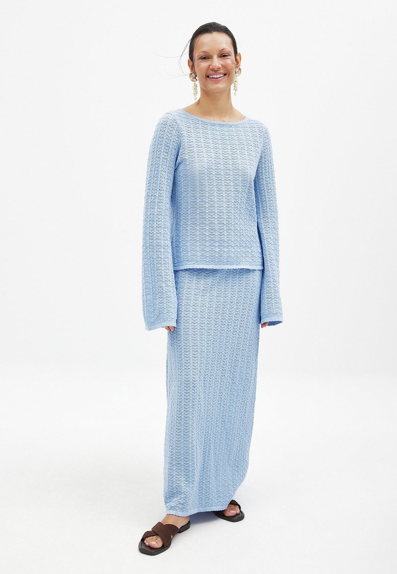 Svetlo modra pletena obleka, ki vključuje dolge rokave in usklajen dolg krilo. Ima teksturirano tkanino z valovitim vzorcem.