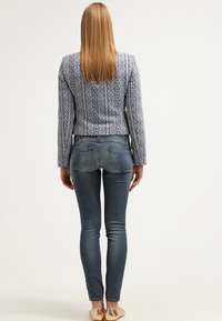 Wzorzysty niebiesko-biały żakiet o dopasowanym kroju, zestawiony z ciemnymi obcisłymi dżinsami i jasnymi balerinami, widziany od tyłu.