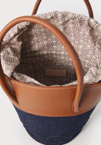 Bucket bag with brown leather handles and denim body. Interior features a patterned fabric lining and a small brand tag.