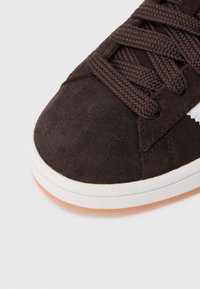 Sapatilha castanha de camurça com parte superior texturizada, atacadores castanhos planos e sola de borracha branca. Contraste de cor distinto na borda da sola.