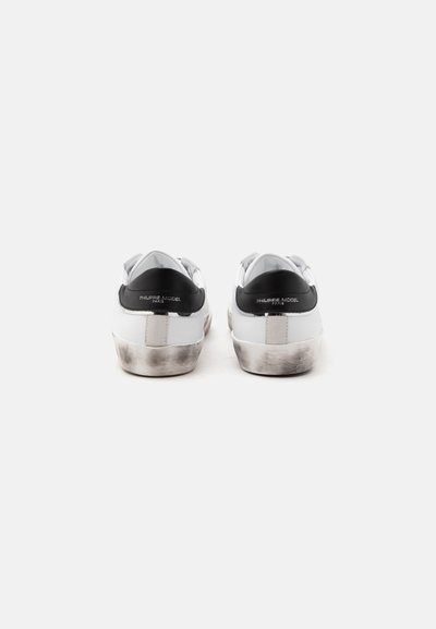 Paire de baskets blanches avec languettes noires au talon et semelles légèrement sales, vues de dos sur un fond clair et uni.