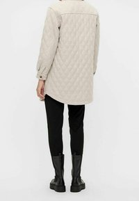 Chaqueta acolchada de color beige claro con un diseño largo, puños abotonados y un cuello plano, combinada con pantalones negros y botas negras hasta el tobillo.