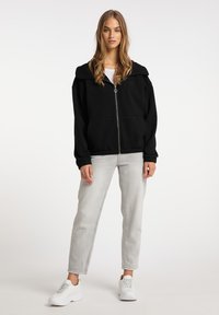 Felpa nera con zip, colletto ampio, polsini a costine e due tasche frontali, abbinata a jeans grigi chiari e sneakers bianche.