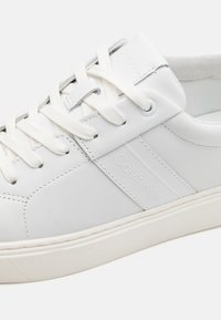Sneaker in pelle bianca con lacci bianchi, punta arrotondata e discreta personalizzazione Calvin Klein sul lato. Texture liscia e design minimalista.