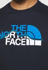 Camiseta de algodón azul marino con un gráfico en blanco y azul del logo "The North Face" y un elemento de diseño ondulado. Construcción de cuello redondo.