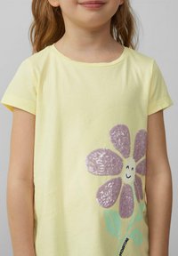 Geel katoenen t-shirt met korte mouwen, versierd met een paarse strass bloem en groene bladeren, met een lachend gezicht in het midden.
