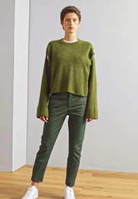 Pull en maille verte à manches longues, associé à un pantalon slim fit vert foncé et des baskets blanches. Sol en bois et mur de couleur neutre en arrière-plan.