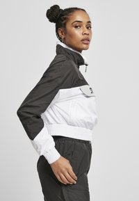 Veste courte en noir et blanc, avec un col montant, une taille élastique et deux poches latérales. Fabriquée en matériau léger et brillant.