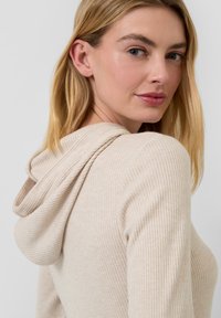 Beige gerippter Hoodie mit weicher Textur, einem lockeren Schnitt und einer drapierten Kapuze. Der Stoff wirkt leicht und angenehm zu tragen.