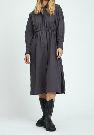 Frau trägt langes dunkelgraues knopfkleid mit geraffter taille, kombiniert mit schwarzen klobigen stiefeln bis zur wadenmitte, steht vor einfarbigem hintergrund.