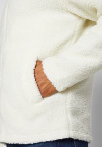 White fleece jacket with a plush texture, featuring a side pocket and a hemline at the waist. The sleeves are slightly loose-fitting.