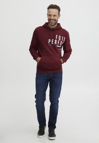 Felpa con cappuccio cremisi e tasca a marsupio frontale, con design grafico testuale, abbinata a jeans blu e sneakers nere.