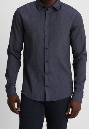 Mørkeblå skjorte med lange ærmer og knapper, med et diskret teksturmønster, båret af en person stående foran en ensartet lys baggrund.