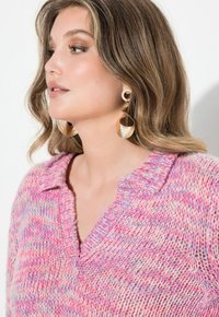 Stickad tröja i rosa och beige med V-ringning och texturerad stickning. Stora örhängen i guldfärg med en spiralformad design.