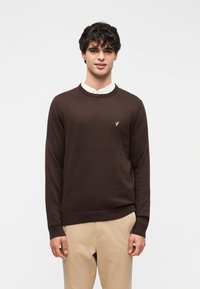 Pull marron tricoté avec un col rond, présentant un petit logo doré sur la poitrine. Associé à un pantalon beige. Texture lisse.