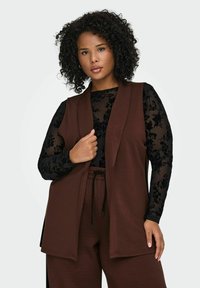 Hnědá vesta bez rukávů vrstvená přes �černý dlouhý rukáv s květinovým vzorem. Vesta má otevřený přední díl a volný střih.