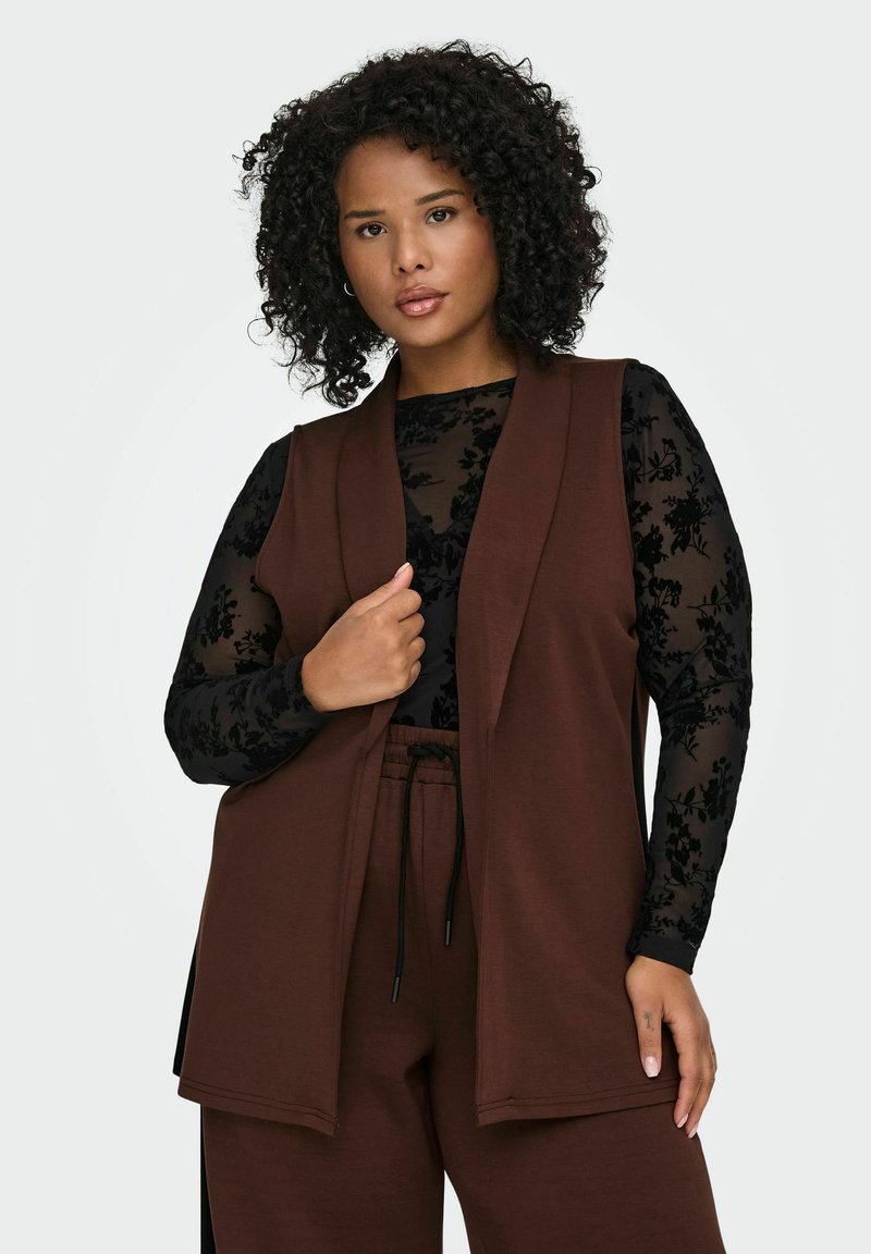 Hnědá vesta bez rukávů vrstvená přes černý dlouhý rukáv s květinovým vzorem. Vesta má otevřený přední díl a volný střih.