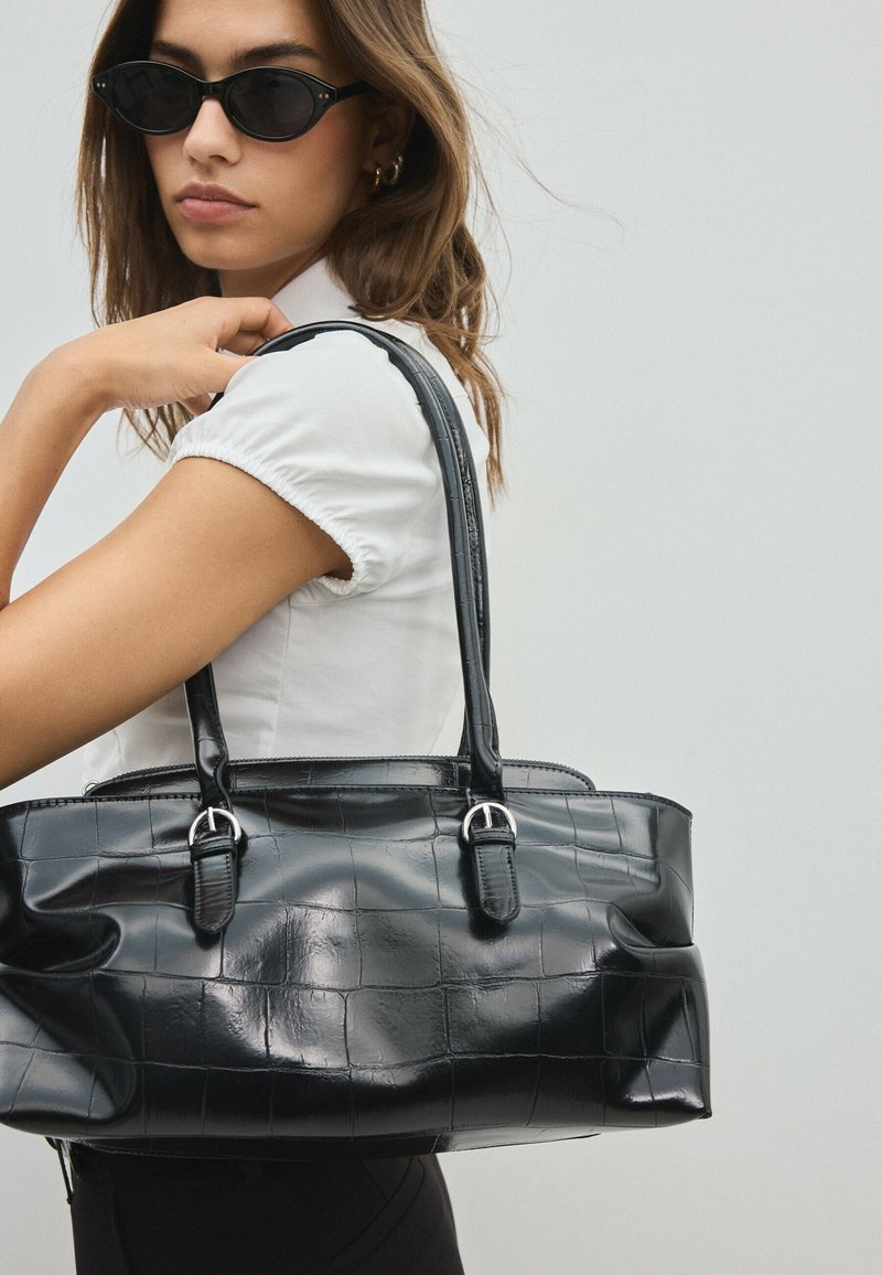 Femme portant des lunettes de soleil noires de forme œil de chat tient un grand sac à main en cuir noir texturé sur l’épaule sur fond gris.